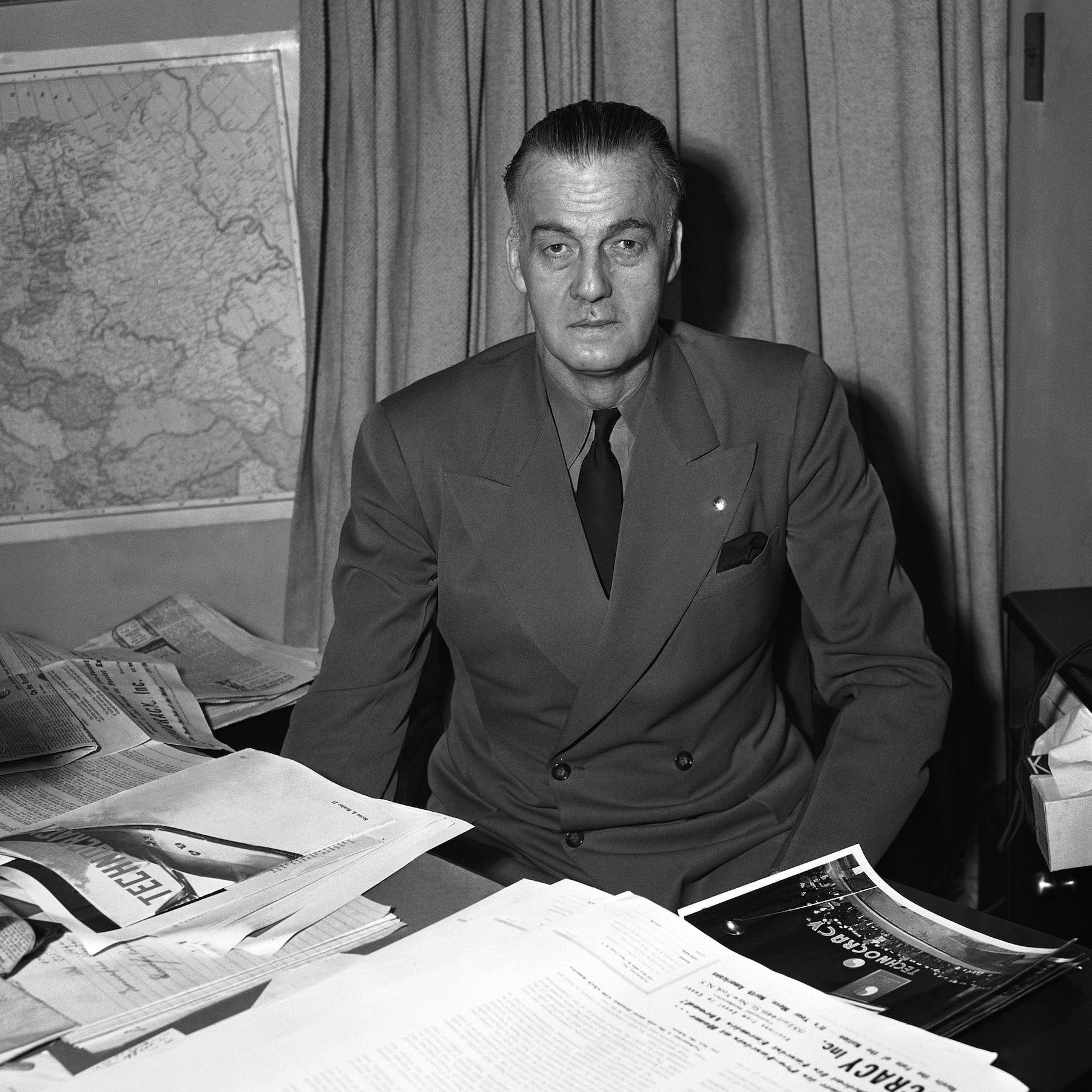 Howard Scott seated at a desk with Technocracy material and a map behind him.