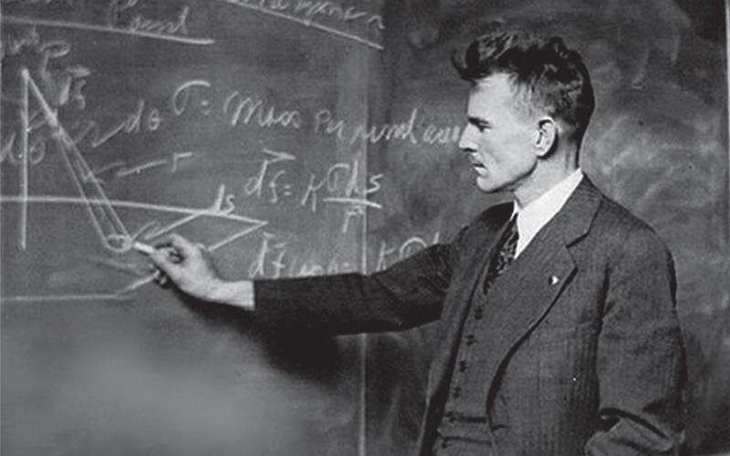 M. King Hubbert standing at a blackboard covered with a technical diagram.